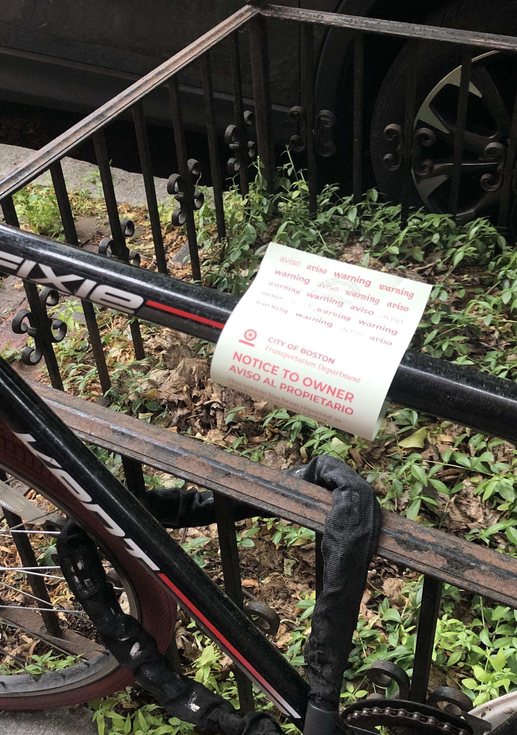 Close-up view of a paper tag attached to a black bicycle. The tag says "NOTICE TO OWNER" and features the City of Boston Transportation Department logo.