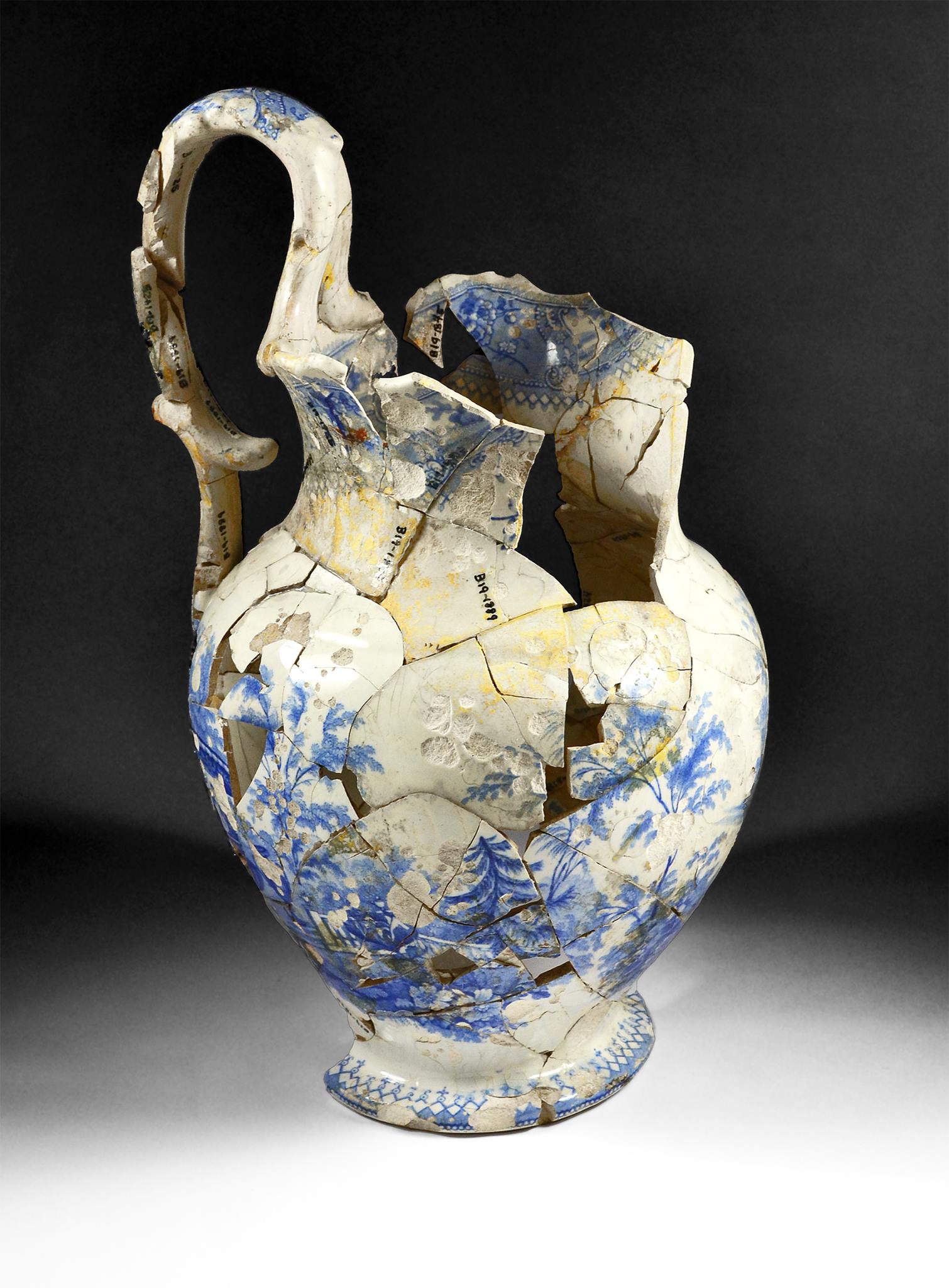 Large ceramic jug recovered from Brook Farm in West Roxbury. It was recovered in pieces by archaeologists and glued back together. It is overall white ceramic with a pale blue printed decoration on the exterior with plants and an urn. It is taller than it is wide, at 12 inches tall and six inches wide with a large handle that sticks up above its rim. The spout of the jug is missing and there are several holes in the body from missing pieces of ceramic