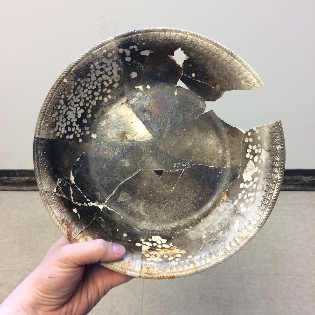 hand holding up a ceramic basin. It is blackened from having been through a fire and has been glued together from multiple fragments. There is a large piece missing from the rim.