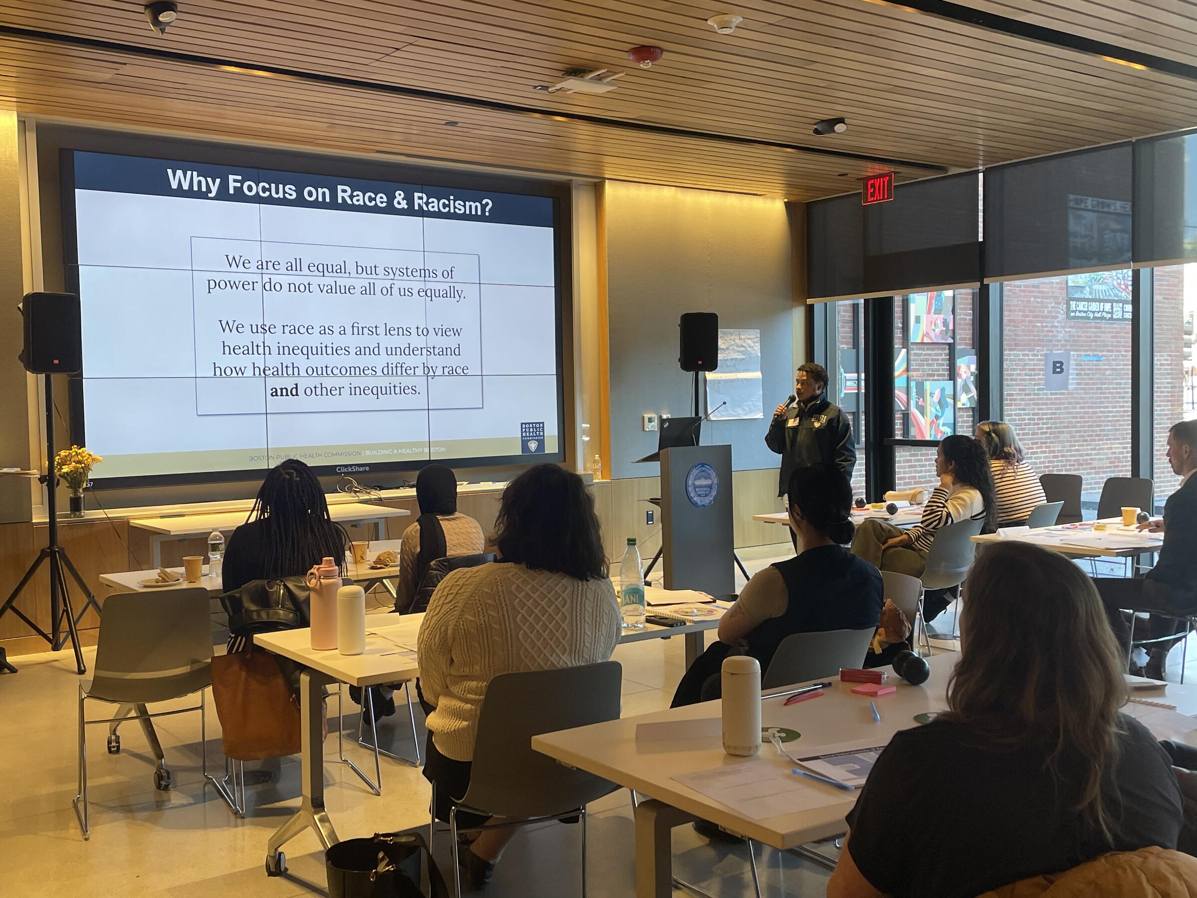 Alex at the front of the room speaking into a microphone, addressing a room of City workers. There is a presentation projected on the wall.