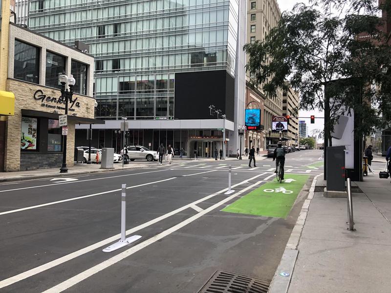 A separated bike lane runs adjacent to a sidewalk in a busy downtown street with mid-rise buildings. A person on a bike is paused in the bike lane.
