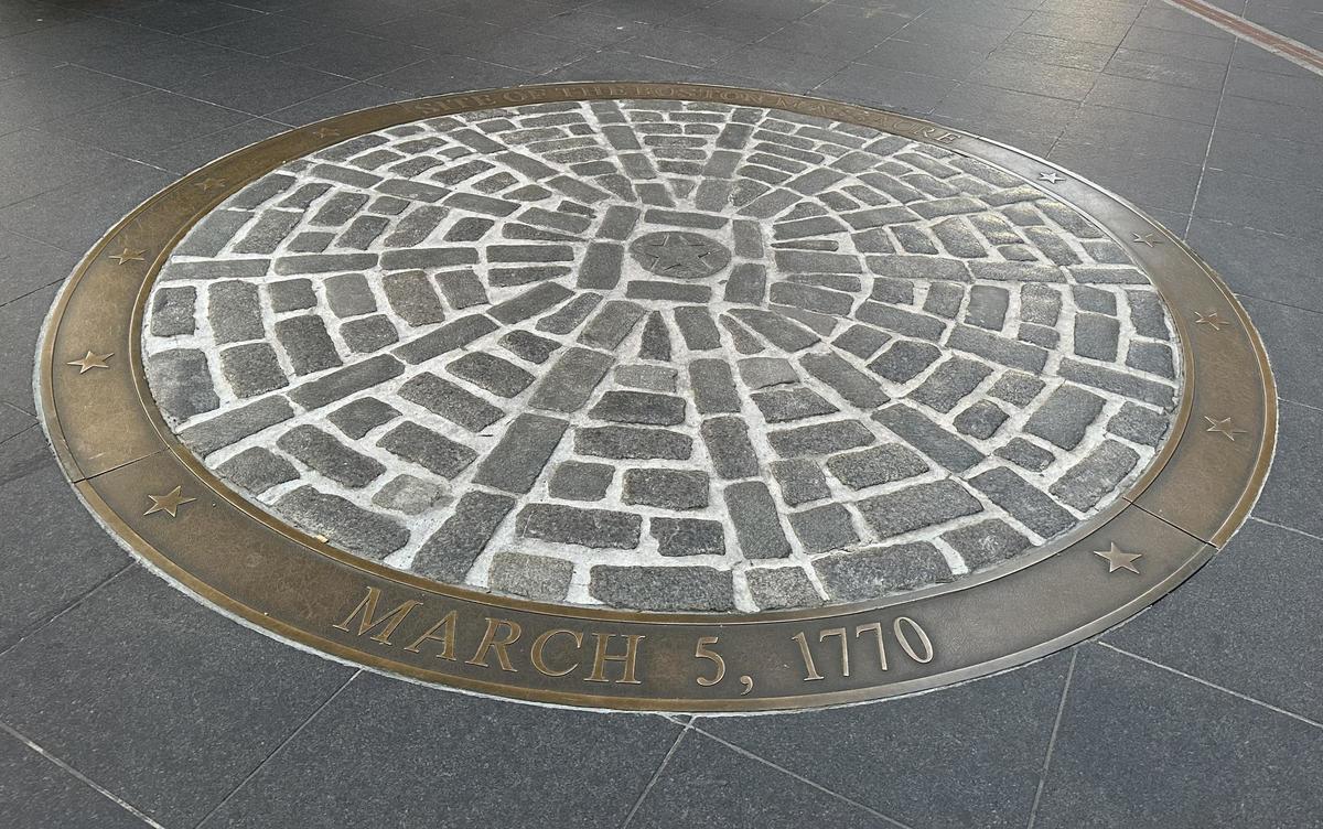 Photo showing the Boston Massacre site marker. It is a circle of pavers embedded in the sidewalk surrounded by a brass ring bearing the text "Site of the Boston Massacre March 5, 1770" 