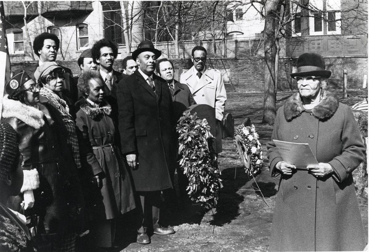 Black and white photograph of Melnea Cass doing a reading in a graveyard while a group of well-dressed members of the Black community look on.