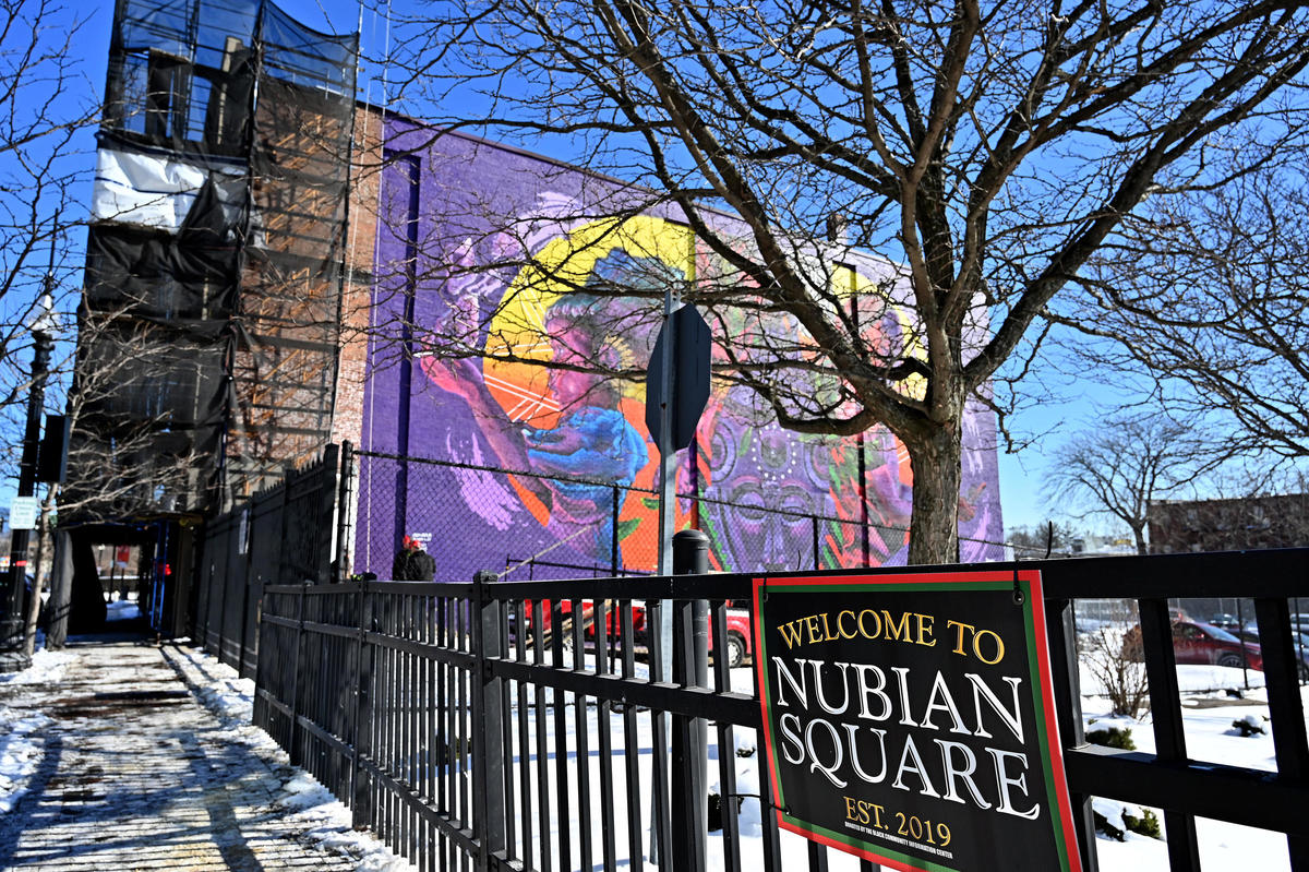 Wintertime view of the Nubian Square sign attached to a fence with a colorful mural in the background showing a little girl and an old woman on either side of a traditional African mask.