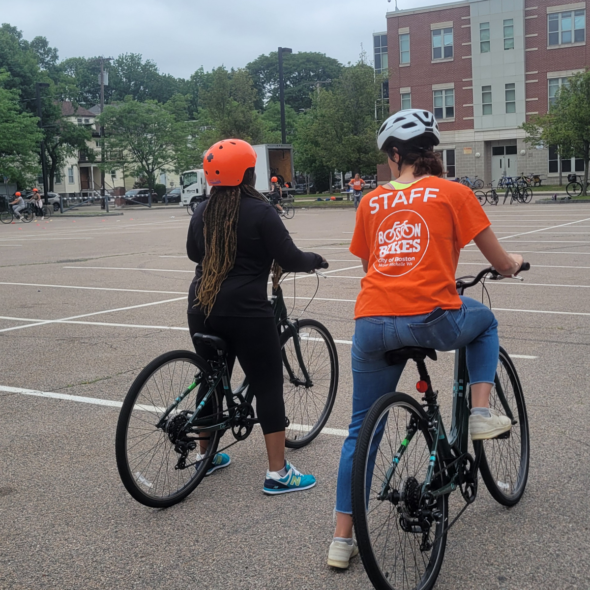 Two people ride their bicycles in a parking lot. One is an instructor, wearing an orange shirt, who is giving some pointers to the other rider