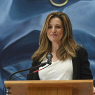 Karin Goodfellow, a white woman with medium-length brown hair, stands at a podium in front of a blue mural.