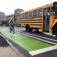 An adult bicycles toward us in a green bike. A yellow school bus is in the adjacent travel lane.