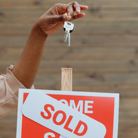  hand holds a set of house keys in front of a sign that says "SOLD" in red letters.