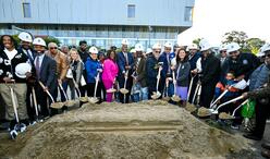 Mayor Michelle Wu with a large group of community leaders, officials, and residents wearing white hard hats stand shoulder to shoulder outdoors, holding shovels filled with sand for a ceremonial groundbreaking. A modern building is visible in the background, and the group includes adults and a child, all facing the camera and smiling.